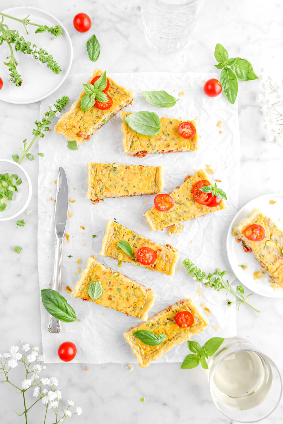 overhead shot of tomato tart slices on parchment paper with basil sprigs and tomatoes around, glass of white wine, and flowers beside