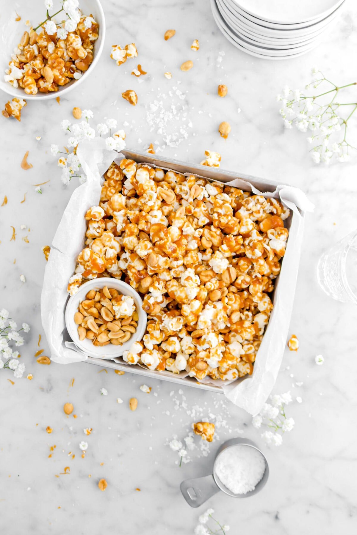 square pan filed with toffee popcorn and a small white bowl of peanuts with flowers around, a small bowl of popcorn beside, and measuring cup of flakes salt