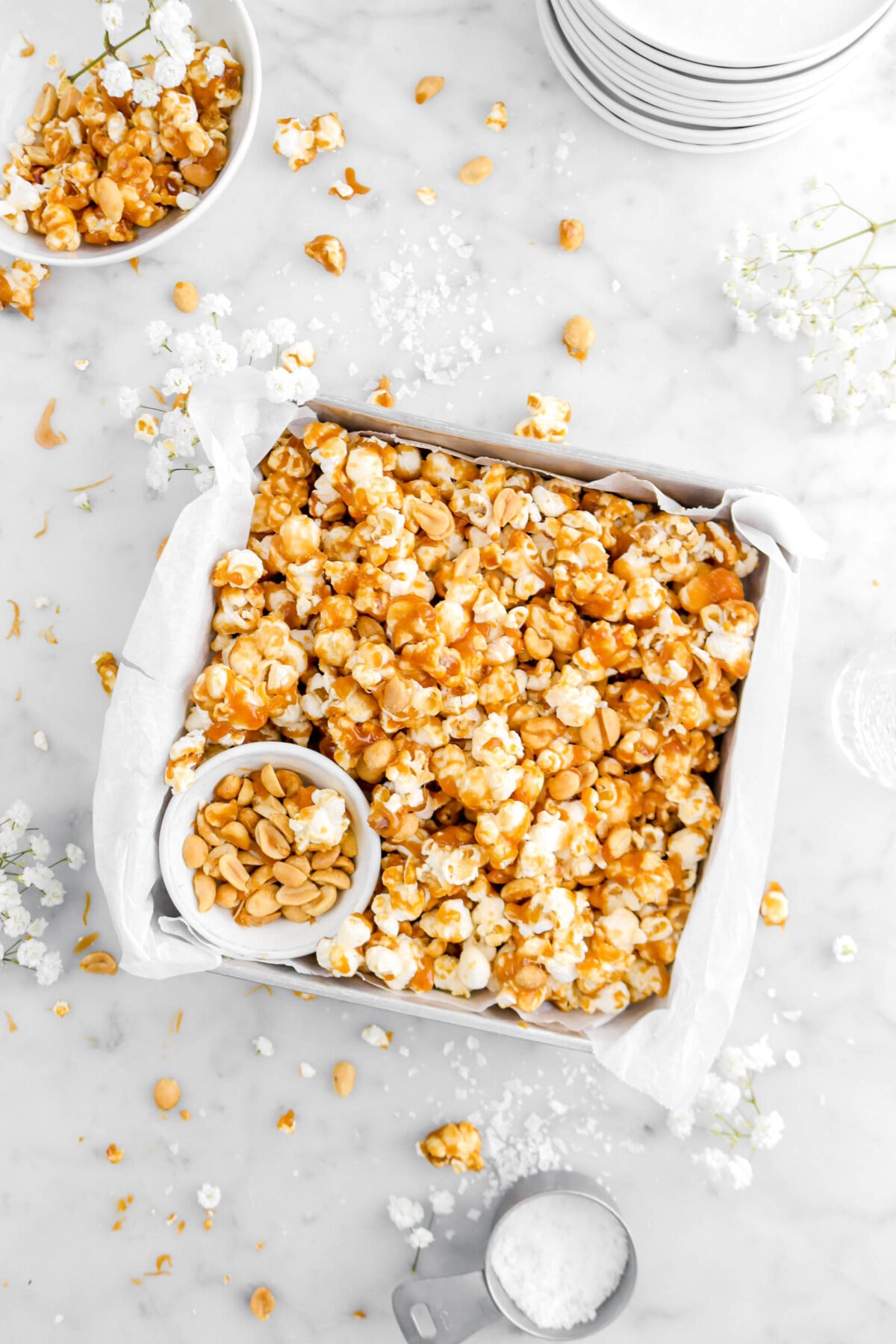 toffee popcorn mix in square pan with bowl of peanuts, peanuts and popcorn scattered around, with flakes salt and small white flowers on marble surface