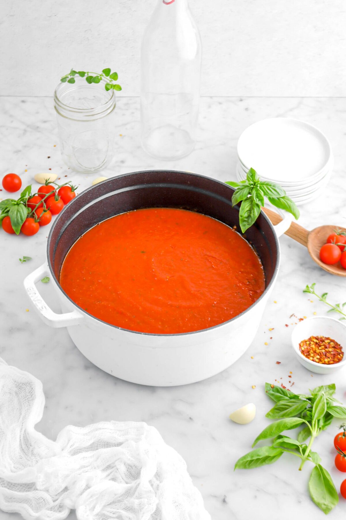 pulled back angled shot of tomato sauce in cast iron pot on marble surface with tomatoes, basil sprigs, bowl of red pepper flakes around, and stack of white plates