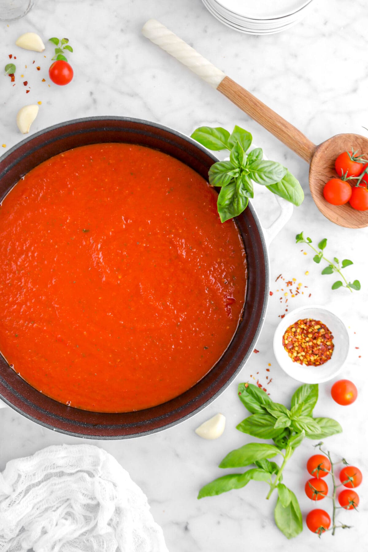 cropped overhead shot of tomato sauce in cast iron pot with basil sprig on handle, wooden spoon beside, herbs, fresh tomatoes, and garlic cloves around on marble surface