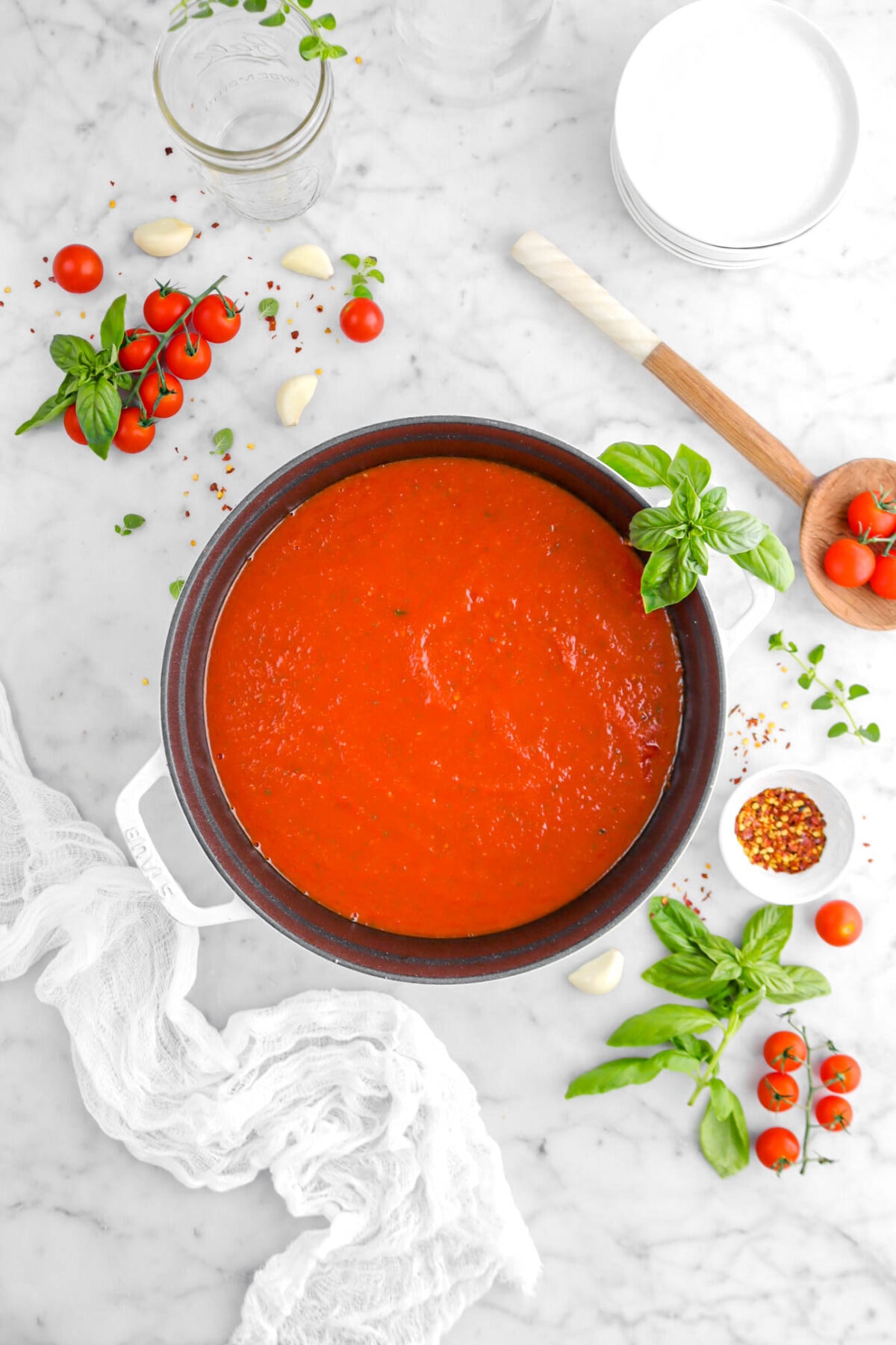 pulled back overhead shot of tomato sauce in large pot with basil and oregano sprigs around, bowl of red pepper flakes, tomatoes on the vine, with white cheese cloth and stack of white plates