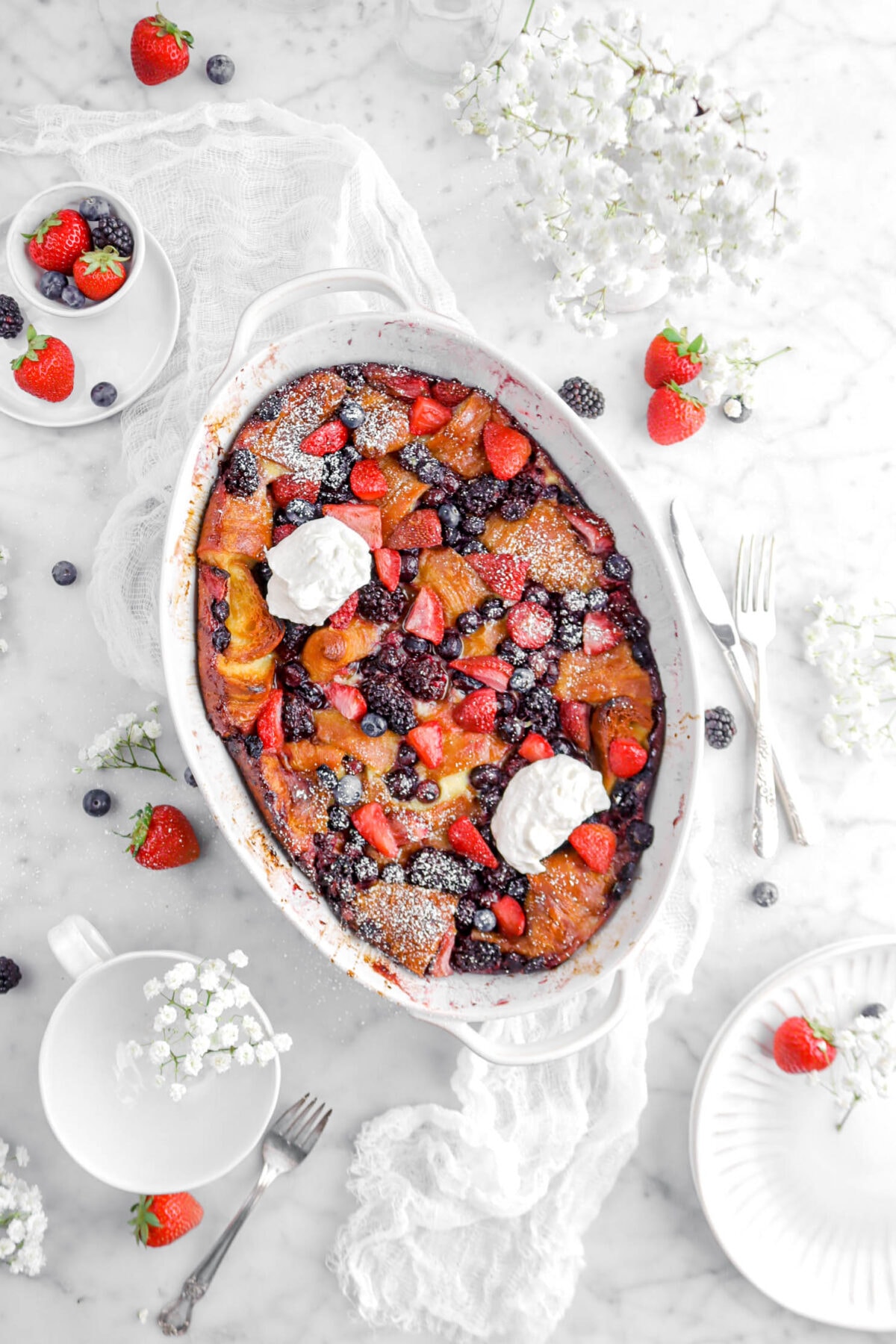 berry bake in white casserole with powdered sugar and whipped cream on top, with white cheese cloth, flowers, whole berries, and plates around
