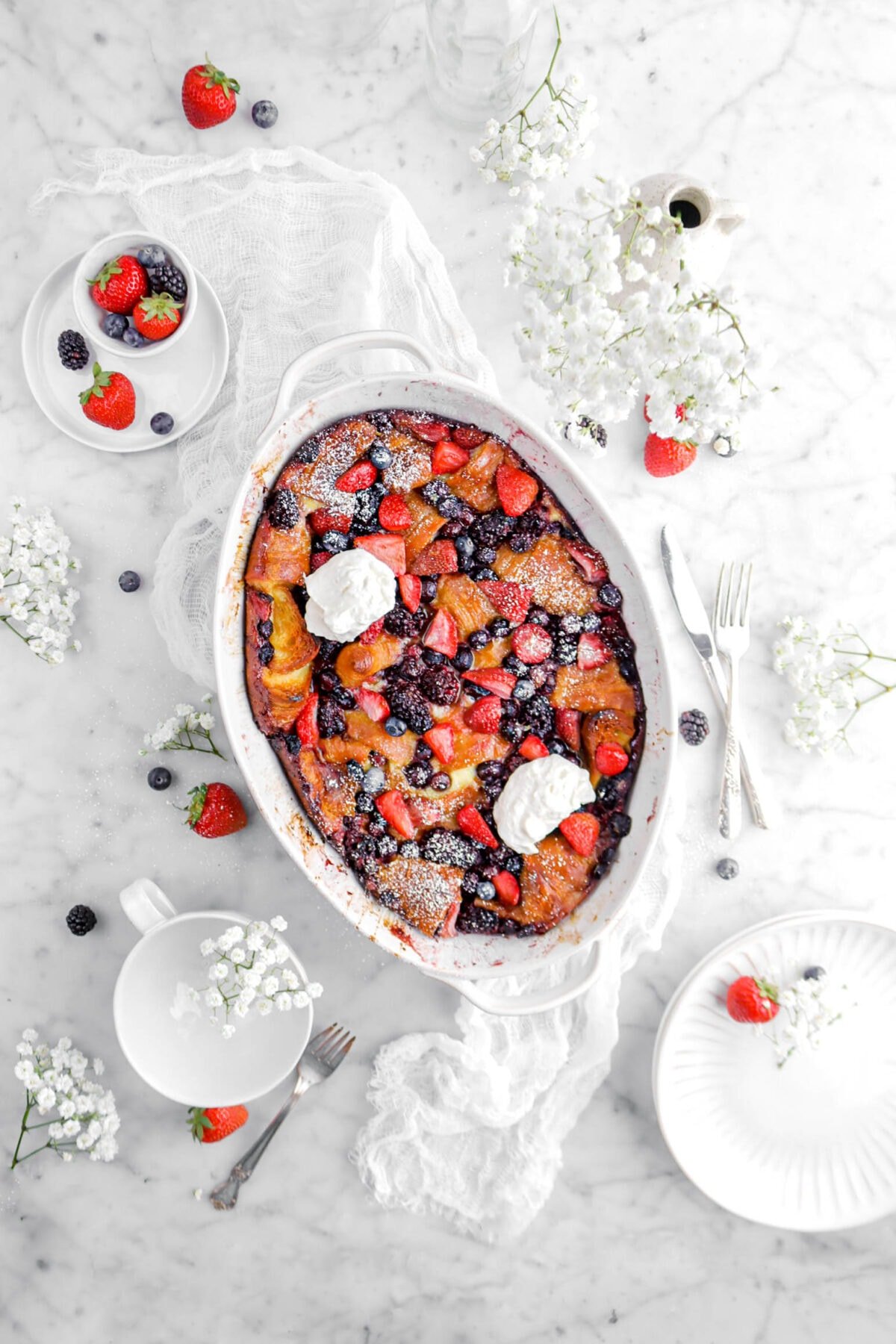 pulled back shot of berry bake in white casserole with flowers, fresh berries, plates, and forks beside