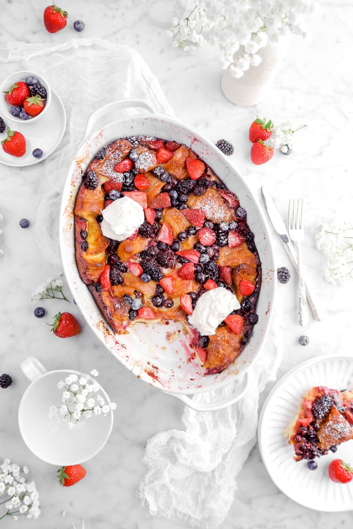overhead shot of triple berry bake on white cheese cloth with slice on white plates beside, fork and knife beside, berries and white flowers around
