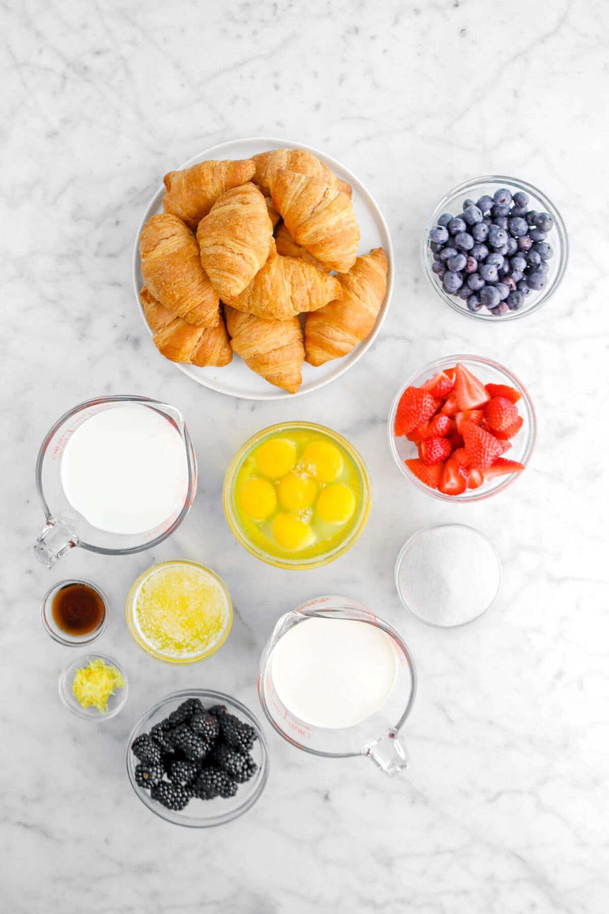 croissants, blueberries, strawberries, eggs, milk, sugar, cream, melted butter, vanilla, lemon zest, and blackberries on marble surface