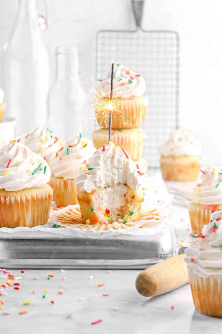 vanilla ice cream filled funfetti cupcake with sparkler burning on upside down sheet pan with more cupcakes around and rainbow sprinkles