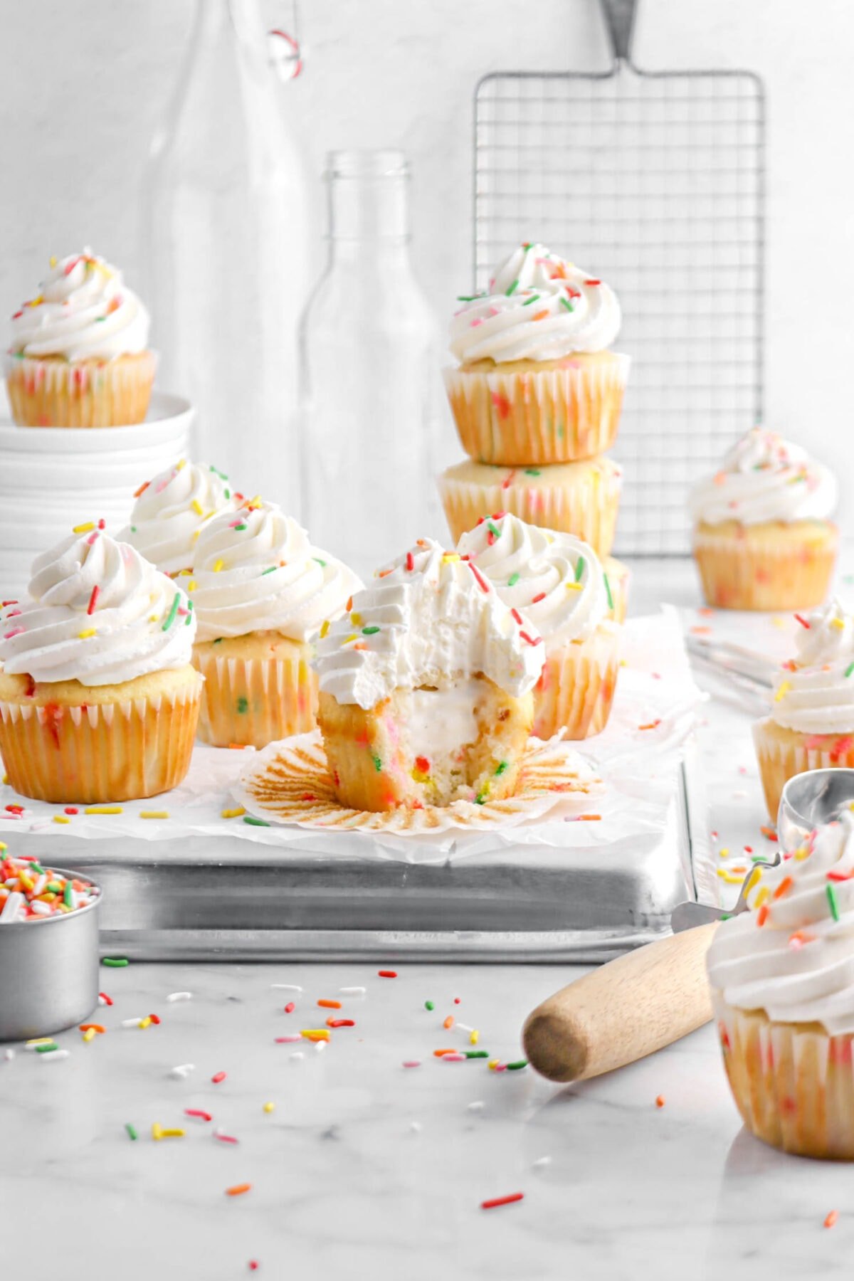 front shot of cupcake with bite missing, eleven more cupcakes around, stack of white plates and empty glasses behind, and rainbow sprinkles around
