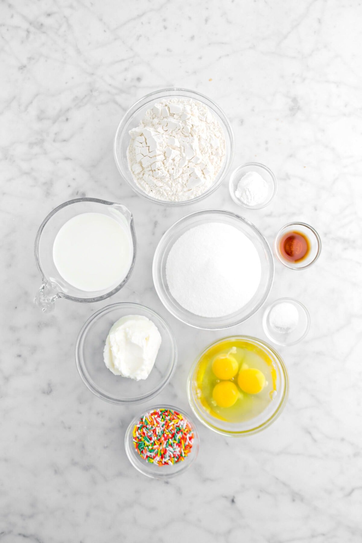 flour, baking powder, vanilla, salt, eggs, sugar, rainbow sprinkles, shortening, and milk on marble surface