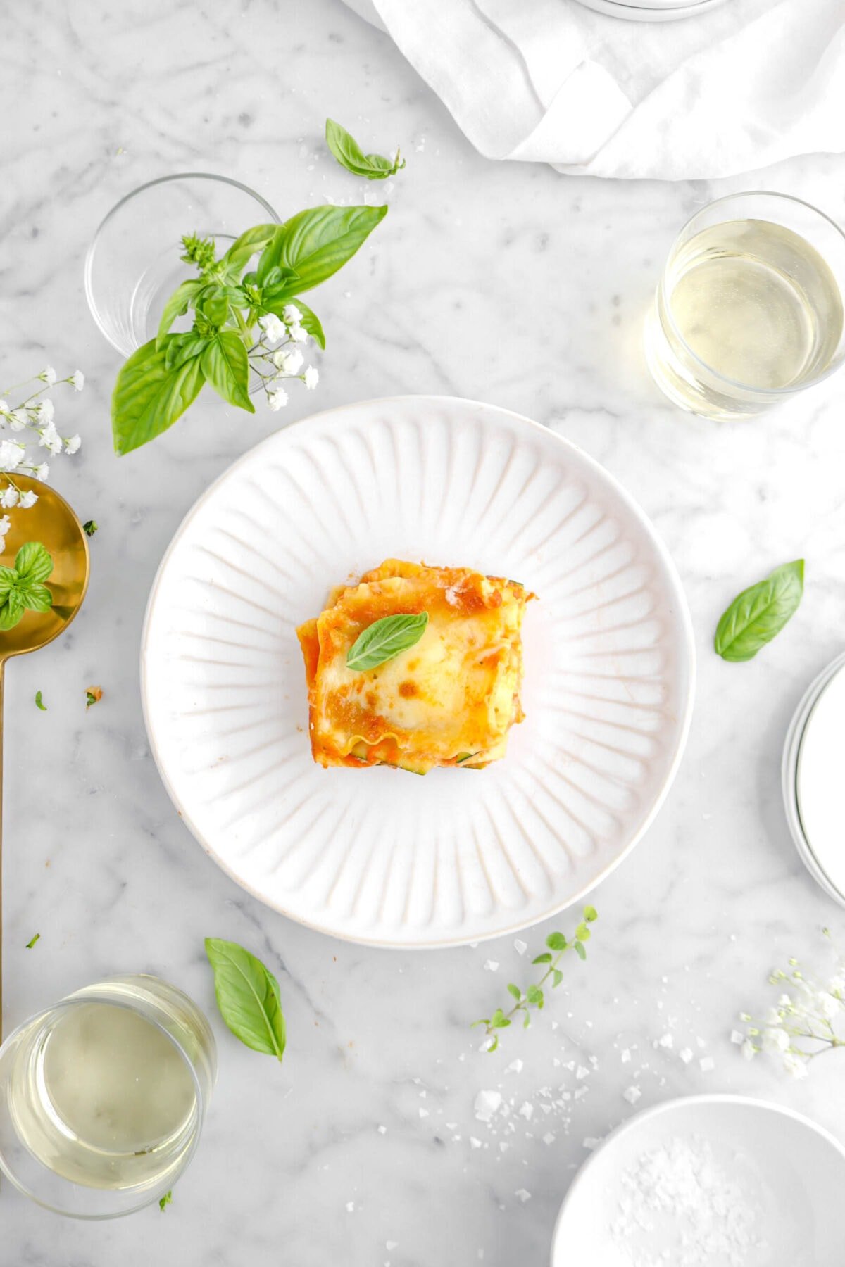 overhead shot of slice of lasagna on white plate with two glasses of white wine beside, herbs around, flaked salt, white napkin, and gold spoon on marble surface
