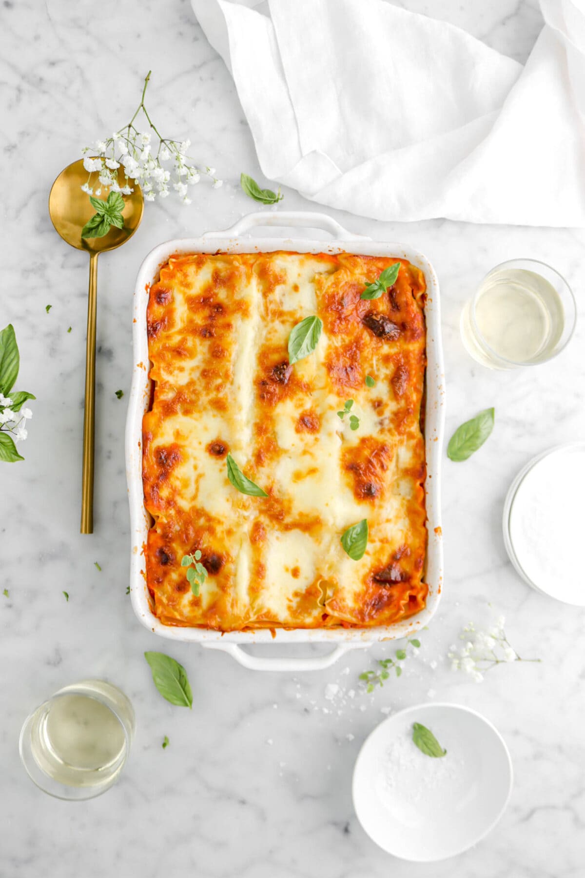 lasagna on marble surface in white casserole with basil and thyme sprigs on top, two glasses of white wine beside, and gold spoon