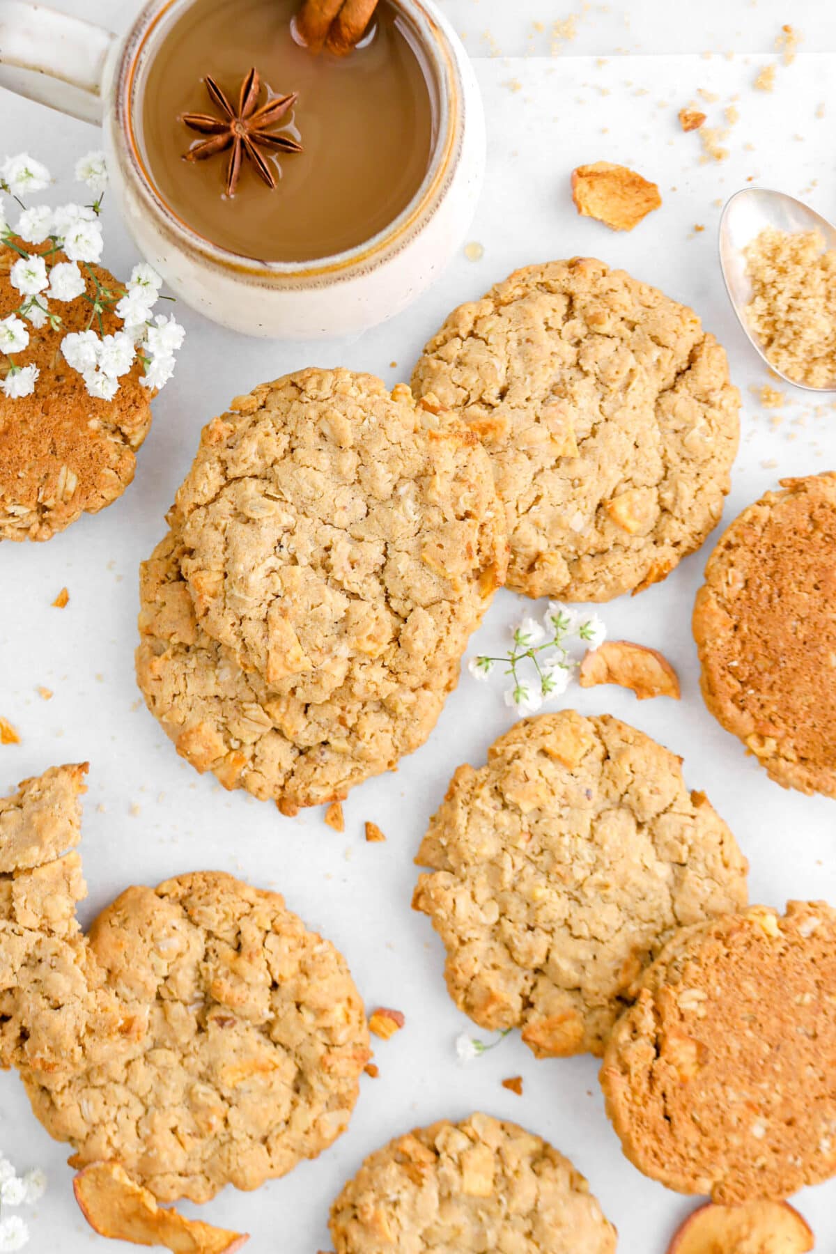 close up of oatmeal cookies on parchment paper with white flowers and mug of cider beside.