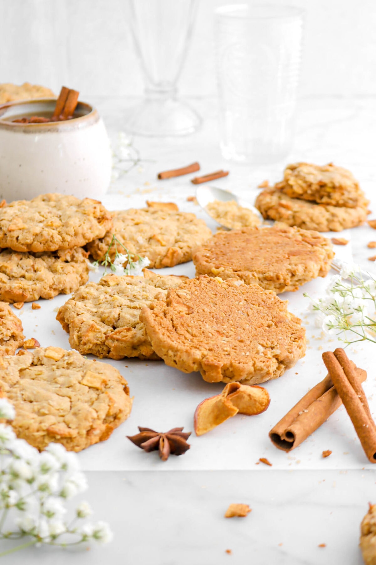 front shot of two cookies leaning against each other, one upside down, with while spices, dried apples, white flowers, and more cookies around on marble surface