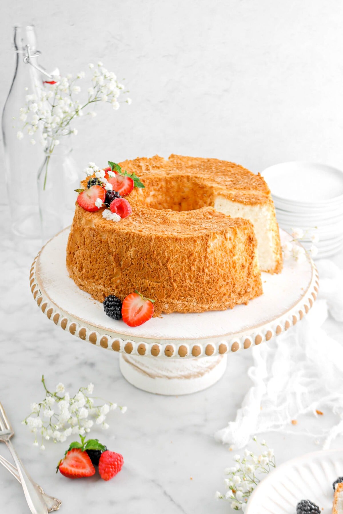 angel food cake with slice missing on white washed cake board with berries on top, fresh mind sprig, and white flowers with cheesecloth beside and forks.