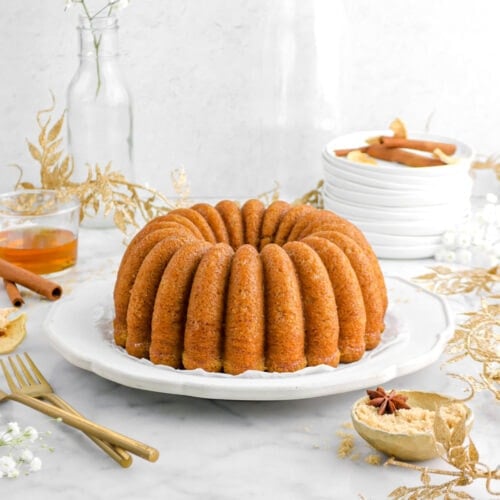 apple bundt cake on plate with gold leaf garand around, two gold forks, and gold bowl of brown sugar beside with glass of bourbon behind.