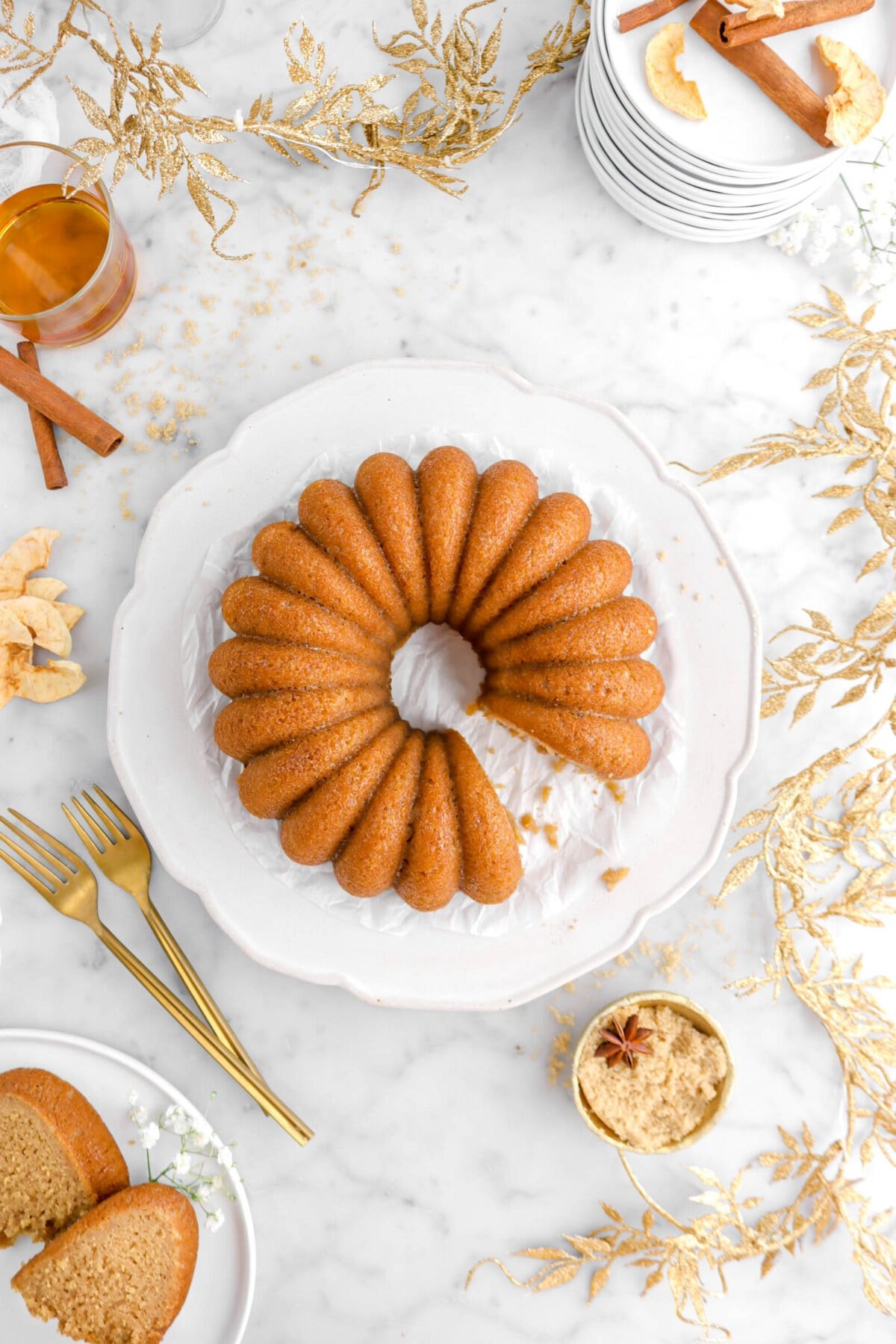 overhead shot of bundt cake with gold leaf garland around, two slices on white plate with white flowers, gold bowl of brown sugar and two forks beside on marble surface