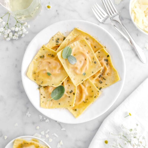 ravioli in white plate with three sage leaves on top with two forks beside and glass of wine beside.