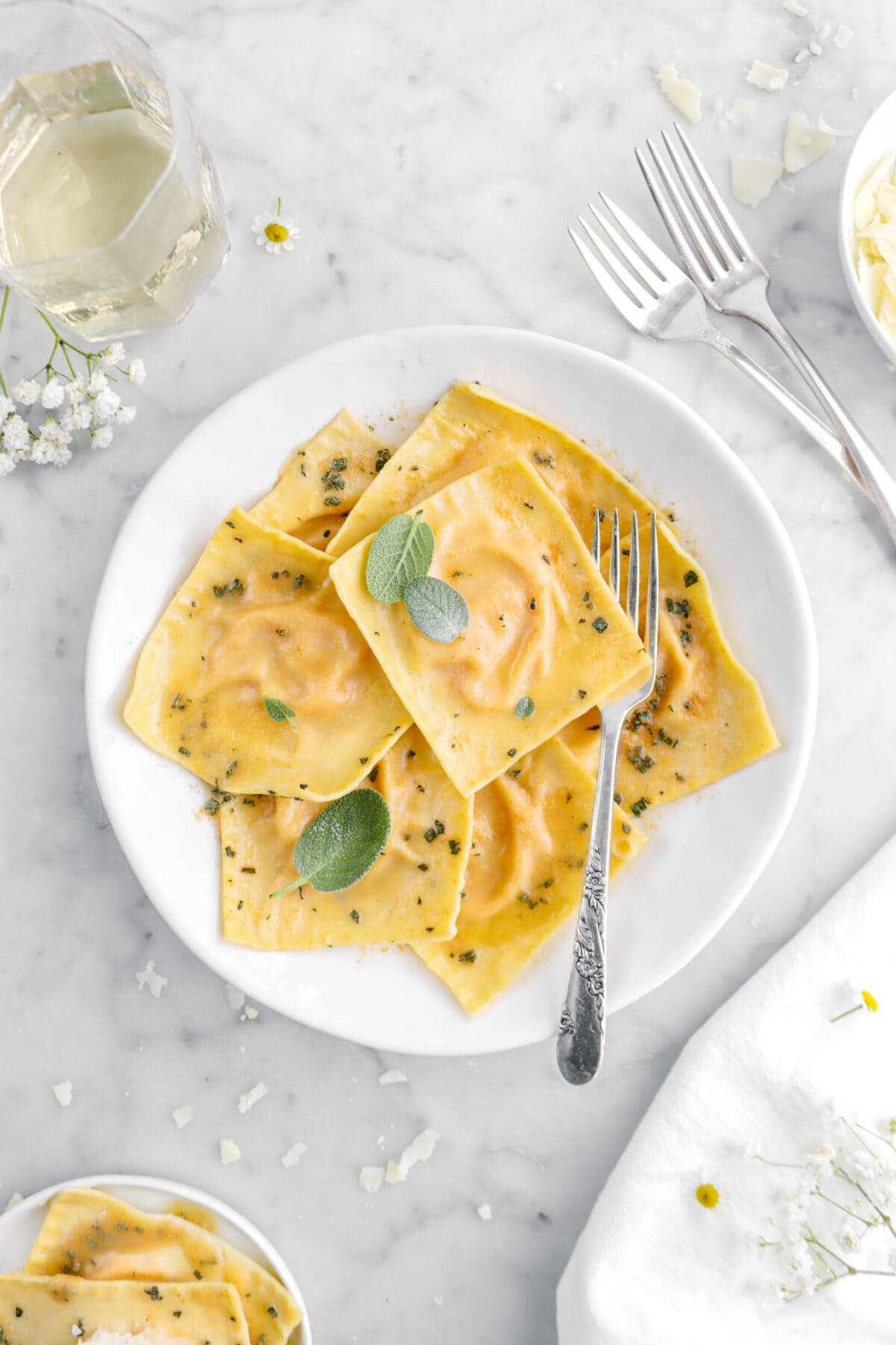 close up of seven on white plate with fork underneath a ravioli, sage leaves on top, with two forks and glass of wine beside.