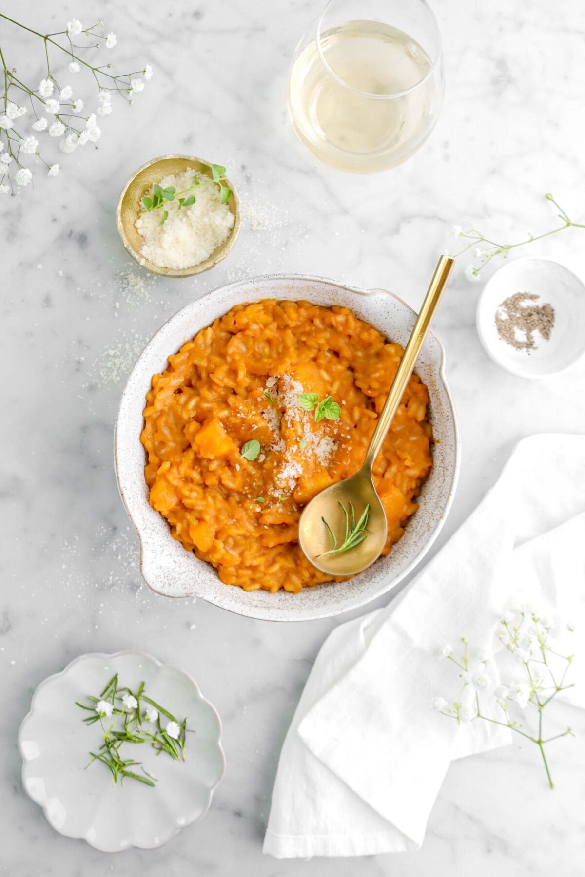 overhead shot of butternut squash risotto on marble surface with two small bowls, a glass of wine, a plate, and white flowers around.