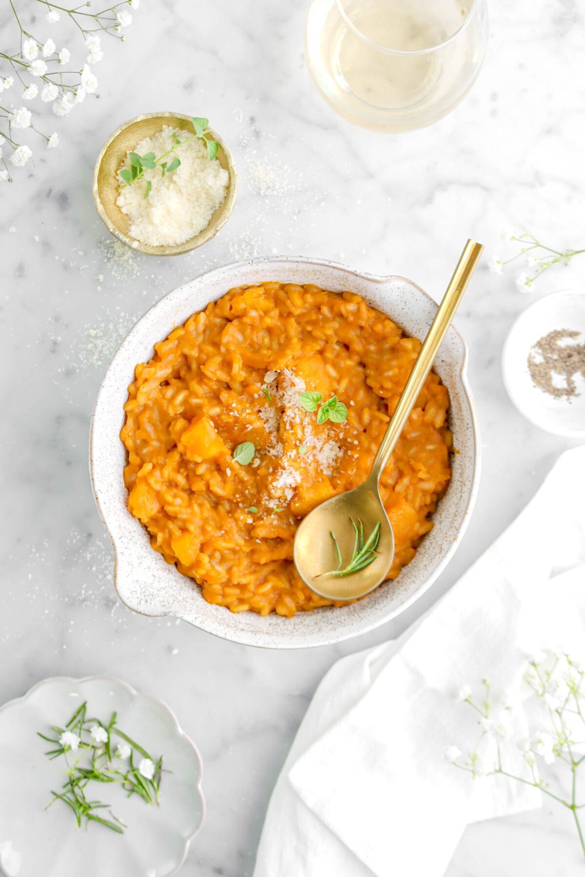butternut squash risotto in bowl with gold spoon on top, a bowl of grated cheese beside, a glass of wine, and white napkin.
