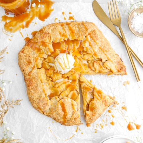 caramel apple galette with scoop of ice cream on top on parchment paper with slice on plate beside, gold leaf garland and jar of caramel to the left side, and fork and knife to the right side