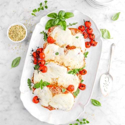 overhead shot of four chicken parmesan on white platter with roasted tomatoes and basil leaves on top, plate of roasted tomatoes, herbs around, bowl of bread crumbs, and stack of white paltes beside on marble surface