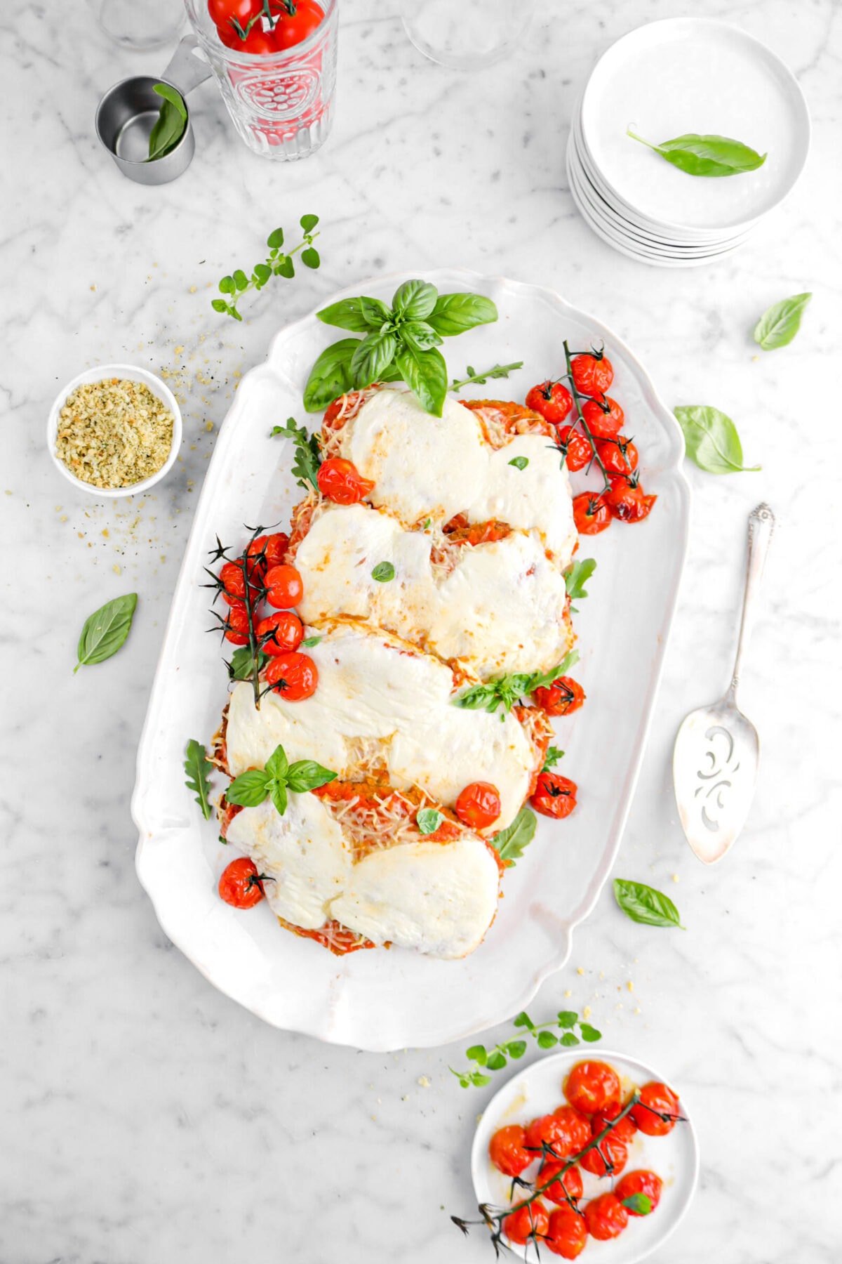 overhead shot of four chicken parmesan on white platter with roasted tomatoes and basil leaves on top, plate of roasted tomatoes, herbs around, bowl of bread crumbs, and stack of white paltes beside on marble surface