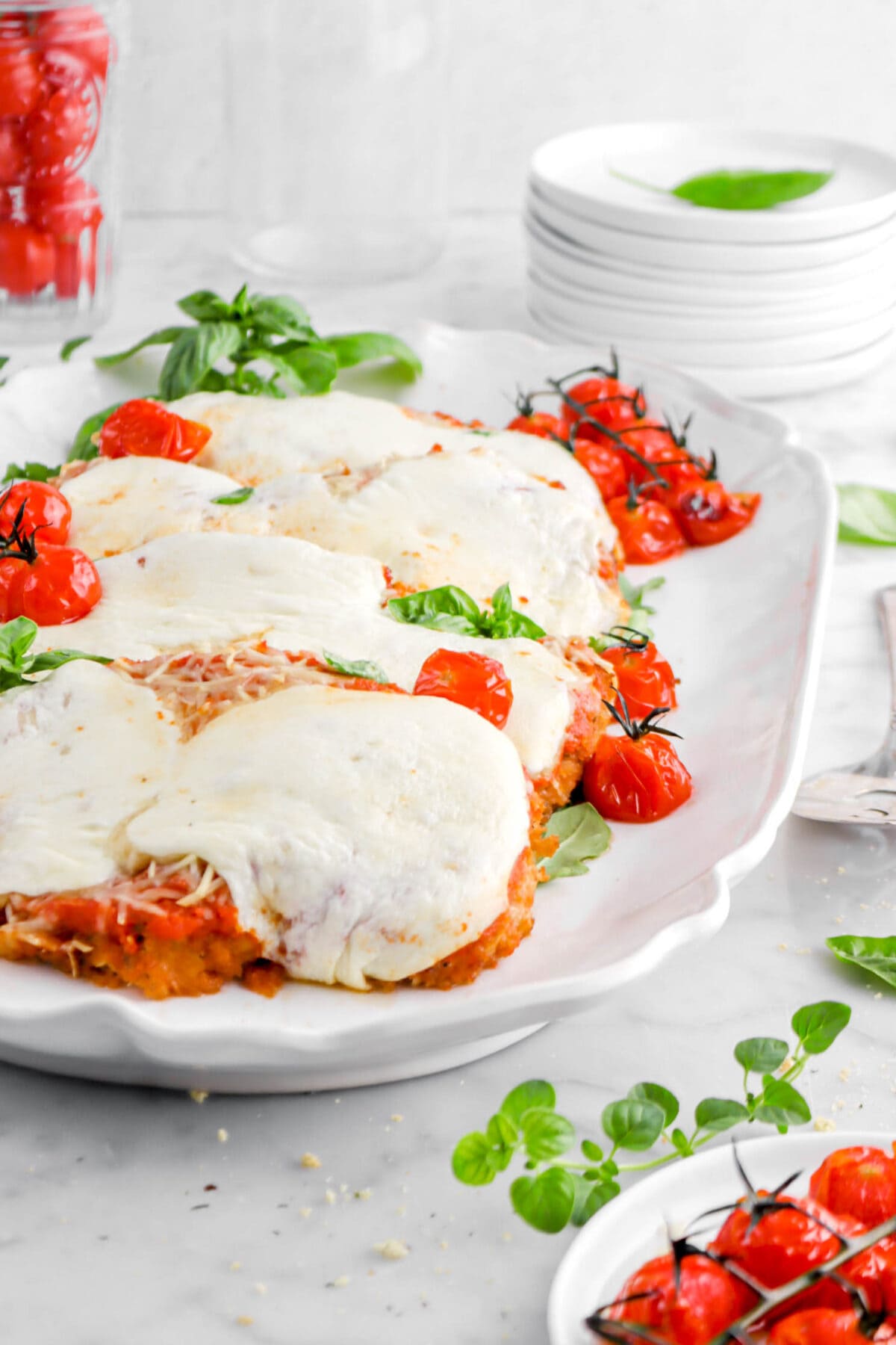 cropped close up of chicken parmesan on white platter with roasted tomatoes on top, stack of plates behind