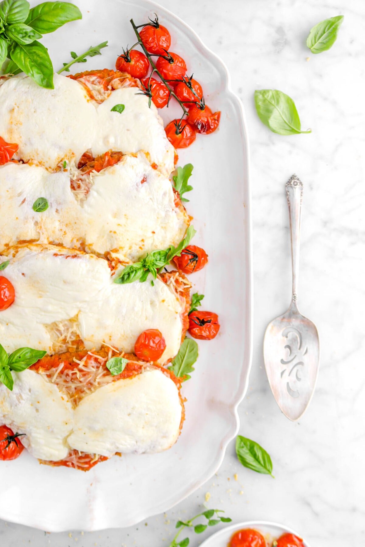 cropped overhead shot of chicken parmesan on white platter with roasted tomatoes and basil leaves on top, and cake knife beside