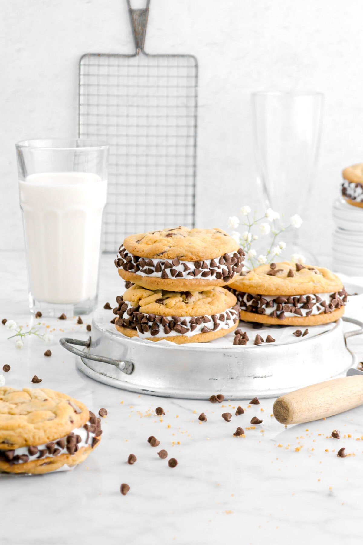 two stacked chocolate chip ice cream sandwich cookies on upside down pie plate with more ice cream sandwiches around, glass of milk, cooling rack, and milkshake glass behind