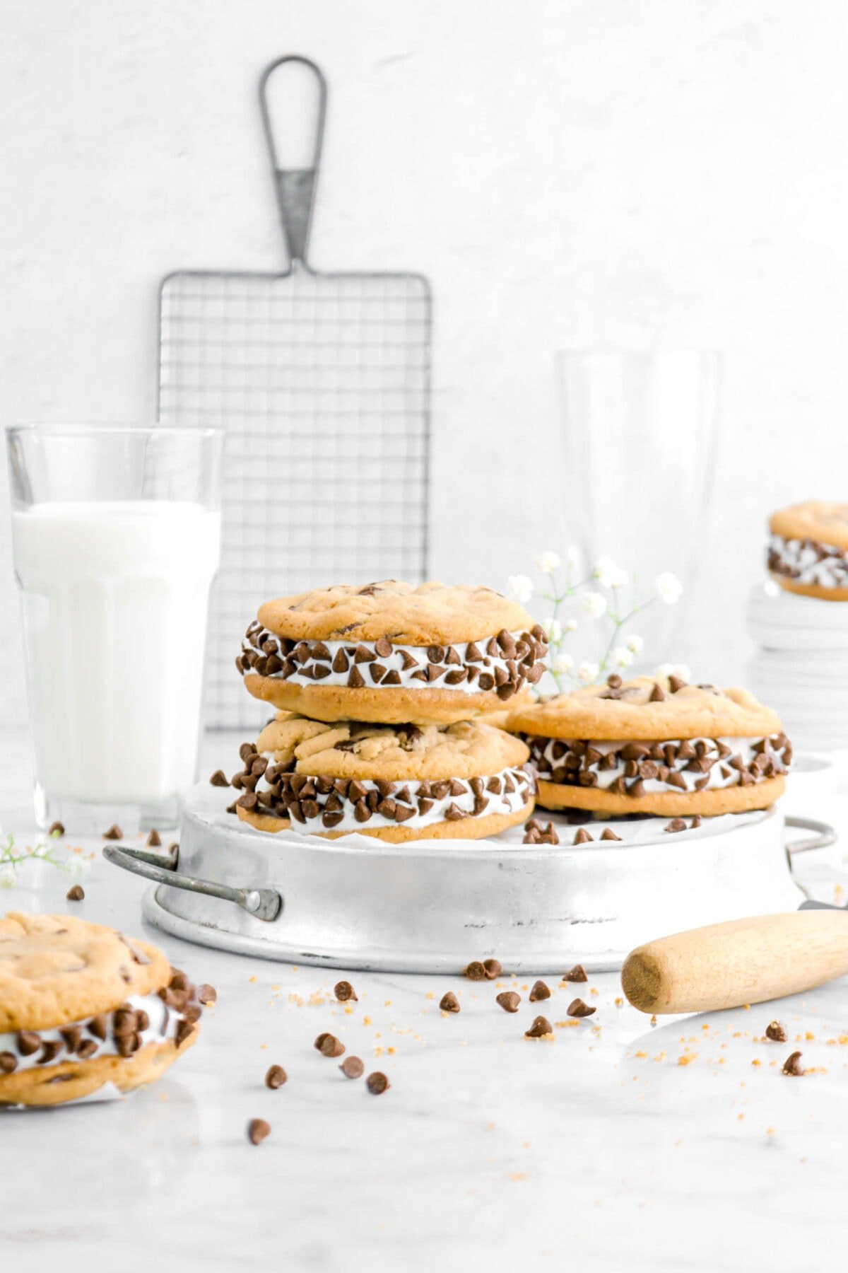 stacked ice cream sanwiches on upside down pie plate with more cookies around, chocolate chips on marble surface with glass of milk and stack of plates behind