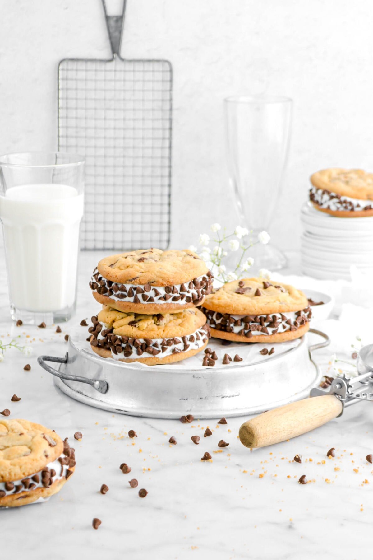 angled shot of stacked ice cream sandwich cookies on pie plate with ice cream scoop beside, glass of milk, white flowers, and chocolate chips scattered around