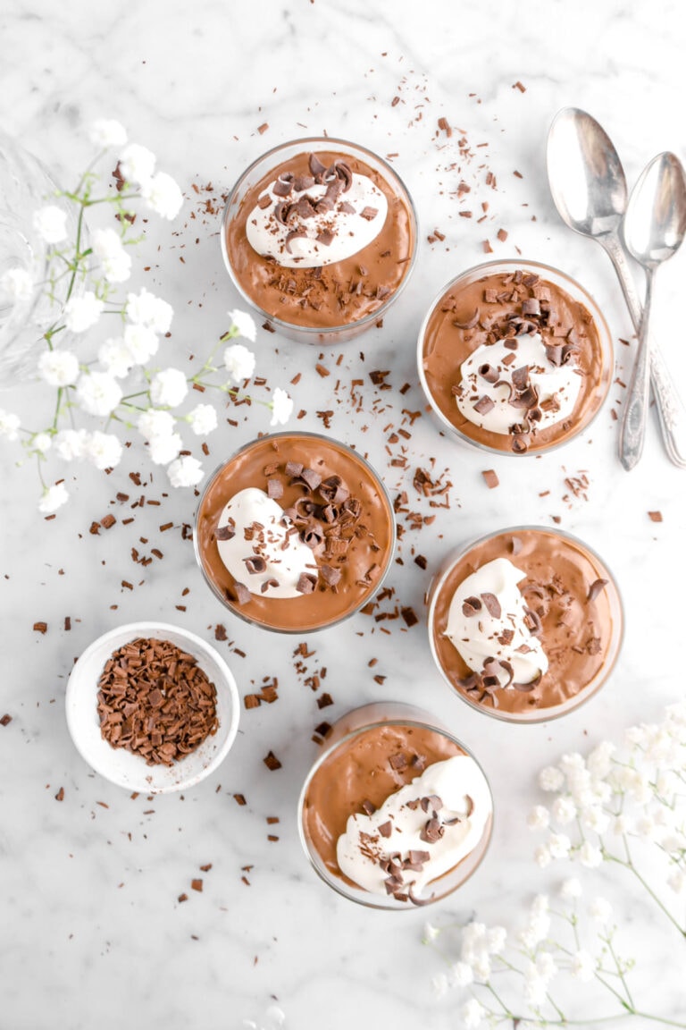 overhead shot of five glasses of chocolate mousse, with bowl of chocolate curls and white flowers around on marble surface