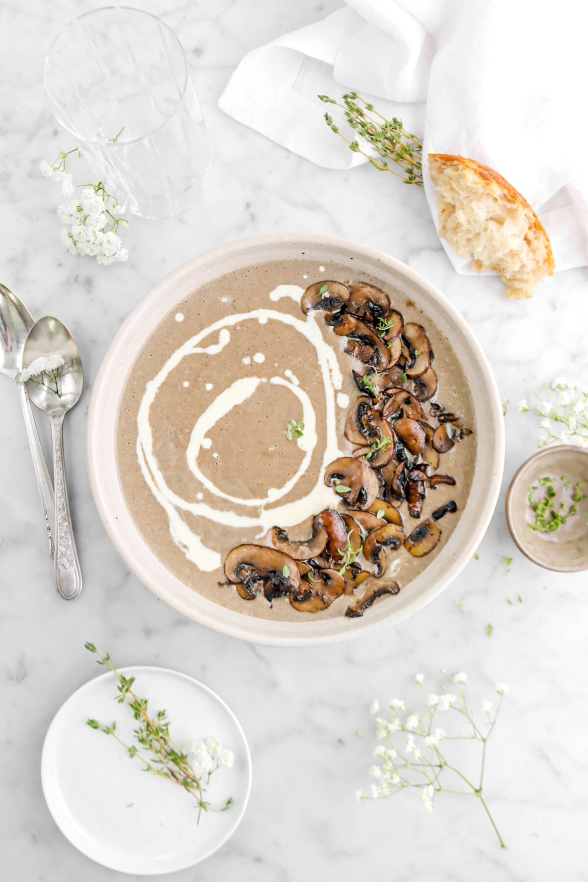 cream of mushroom soup in tan bowl with swirl of cream and mushrooms on top, two spoons, brown bowl, flowers, and thyme sprigs beside on marble surface.