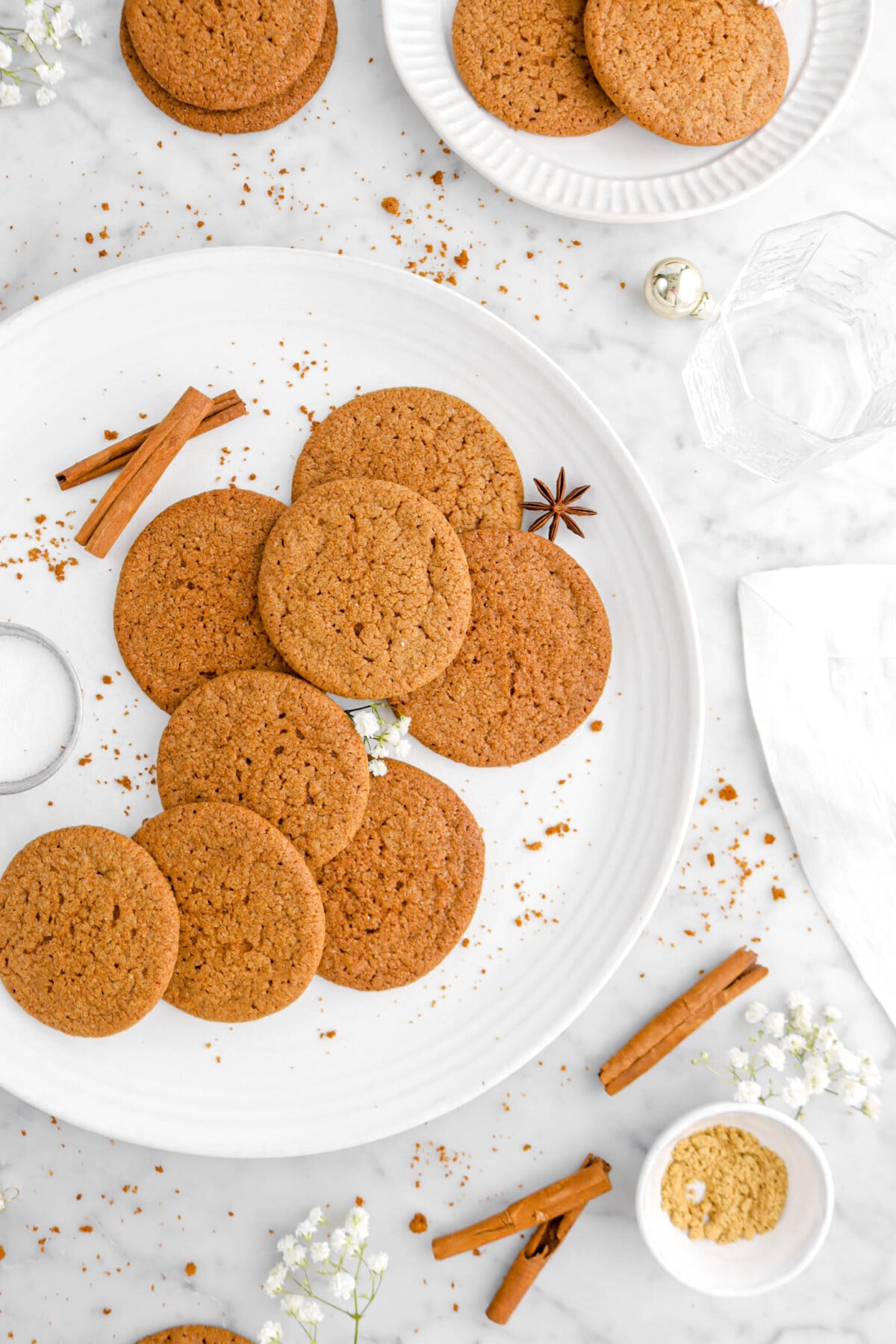 cropped overhead of cookies on plate with star anise pot and cinnamon sticks, white flowers, and more cookies around.