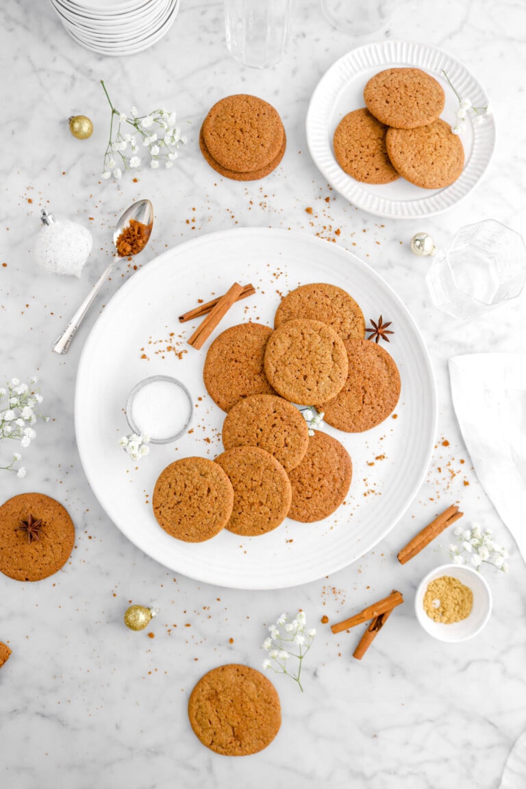 pulled back overhead shot of eight gingersnaps on white platter with seven more around on marble surface with flowers, gold ornaments, cinnamon sticks, and cookie crumbs around on marble surface.