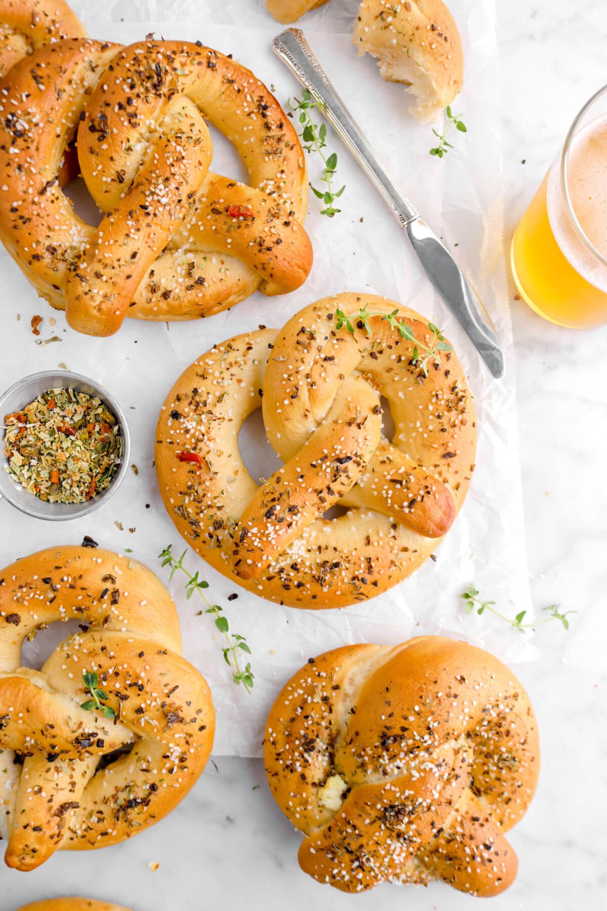 close up of four pretzels with measuring cup of dried herbs beside, glass of beer, a knife, and thyme sprigs around.