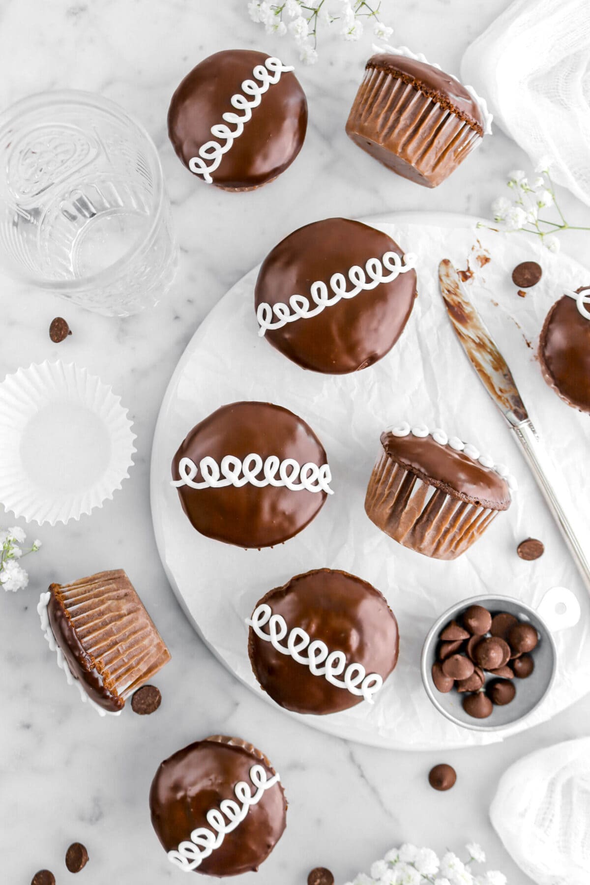 five chocolate cupcakes on upside down plate with one turned on it's side, a knife and measuring cup of chocolate chips beside, with four more cupcakes around on marble surface.