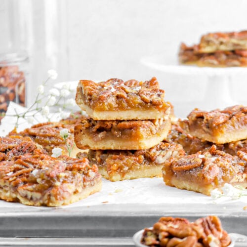 stacked pecan pie bars on sheet pan with more around, white flowers, and measuring cup of pecans on marble surface