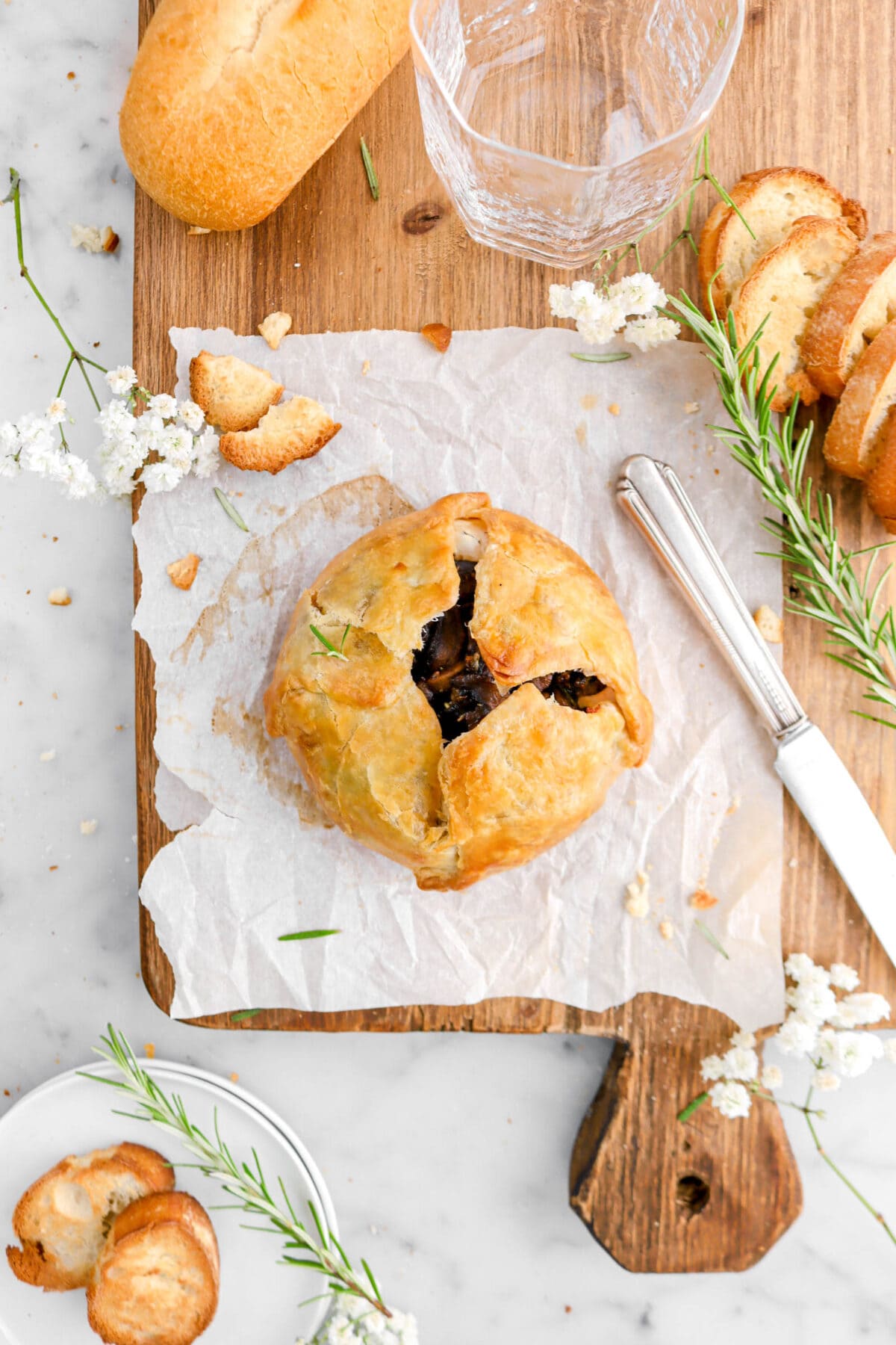 baked brie wrapped in puff pastry on square piece of parchment on wood surface with knife, rosemary sprigs, white flowers, and toasted bread slices around.