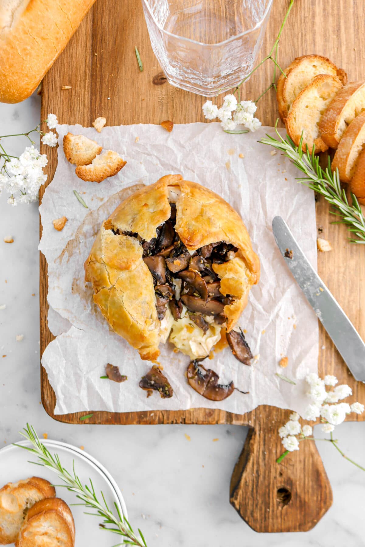 overhead shot of baked brie wrapped in puff pastry with mushrooms spilling out on square piece of parchment paper with flowers, rosemary sprigs, and toasted bread slices beside.