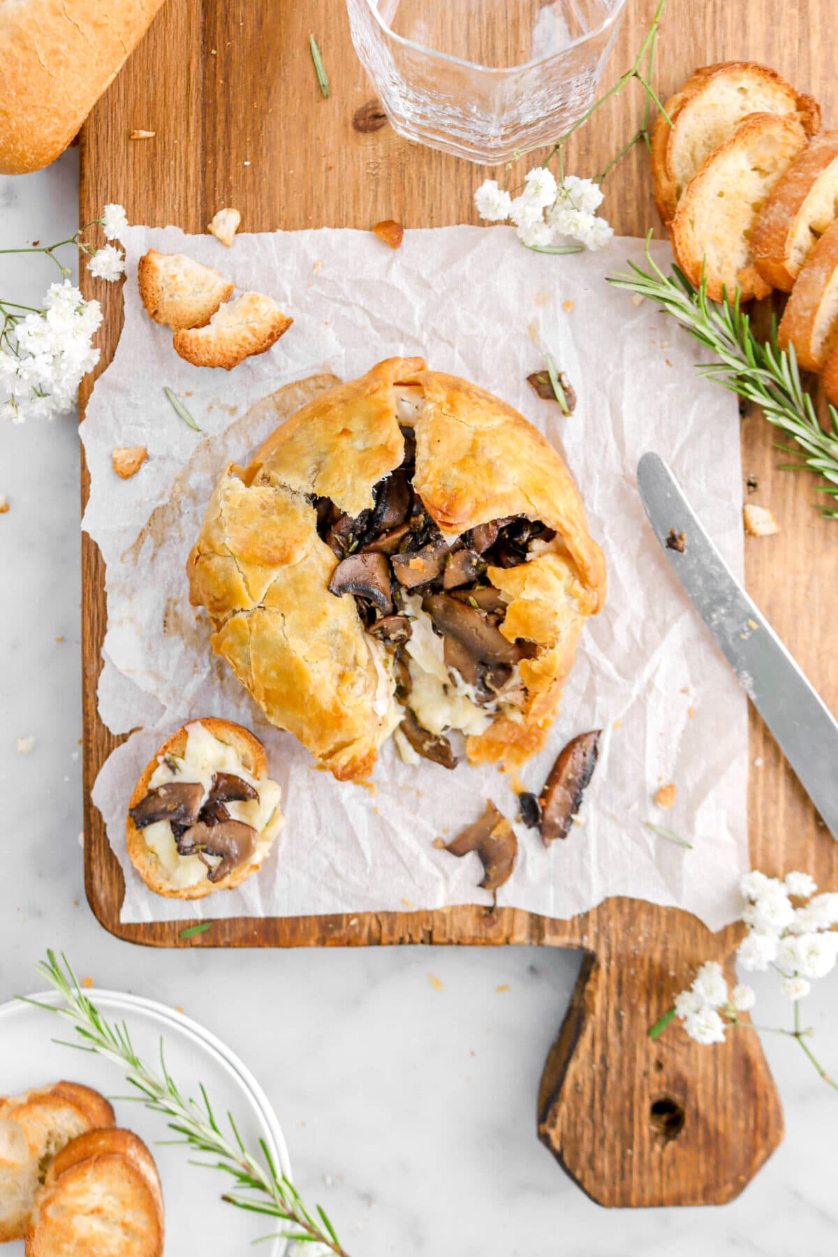puff pastry wrapped brie with mushrooms on parchment paper on wood board with piece of bread with brie and mushrooms on top beside, rosemary sprigs, more pieces of bread, and a knife beside.
