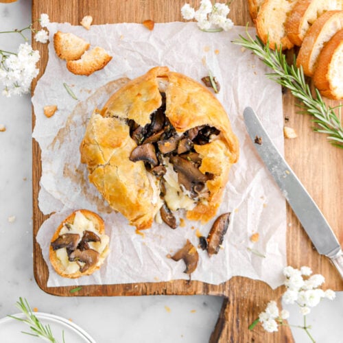 baked brie spilling out of puff pastry with piece of bread beside on parchment paper, white flowers, a knife, and rosemary sprigs around.
