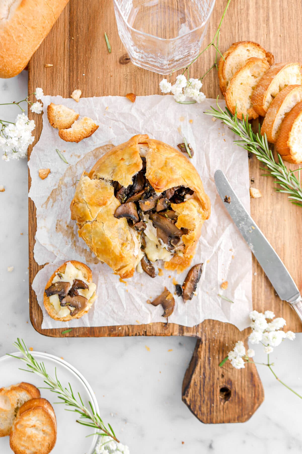 baked brie spilling out of puff pastry with piece of bread beside on parchment paper, white flowers, a knife, and rosemary sprigs around.