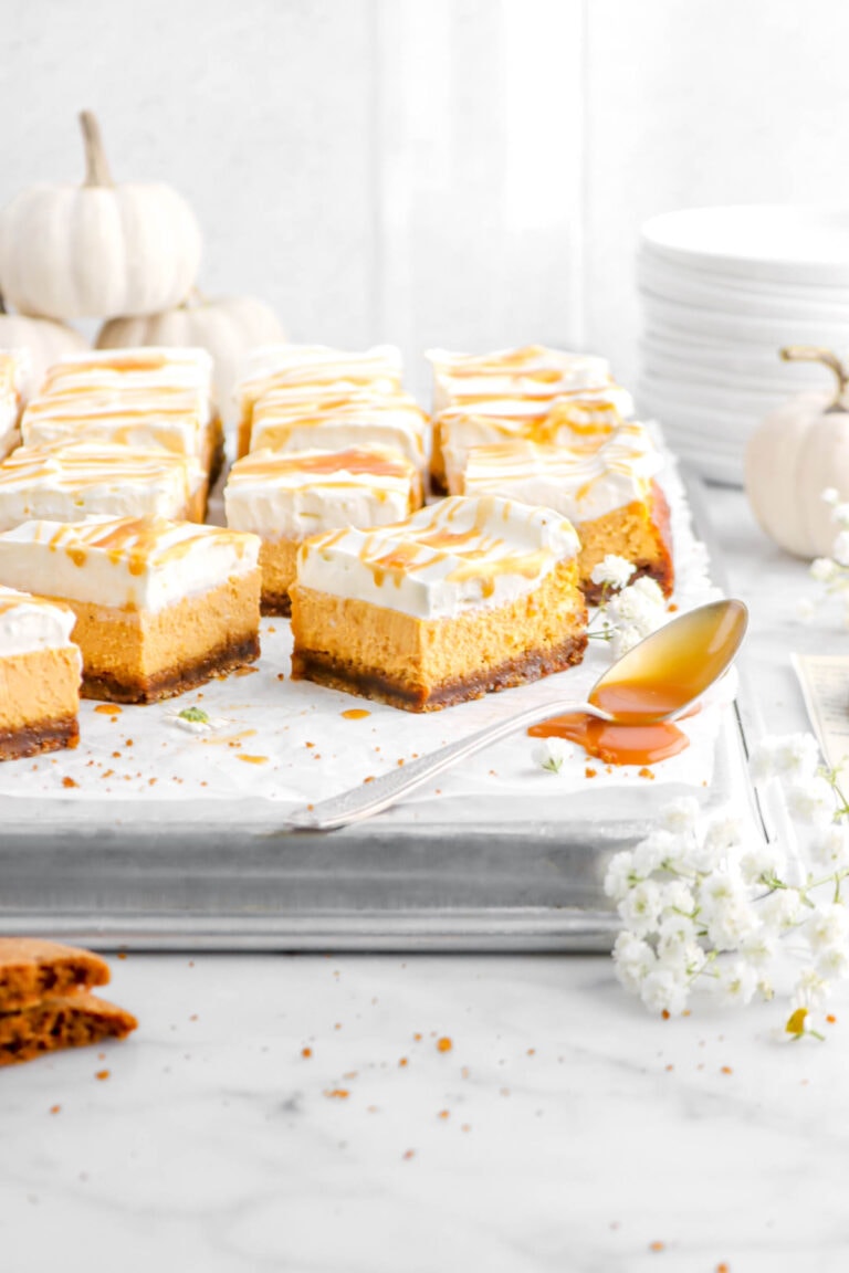 ten pumpkin cheesecake bars on upside down sheet pan with spoon of caramel sauce beside, white pumpkins behind, and stack of plates.