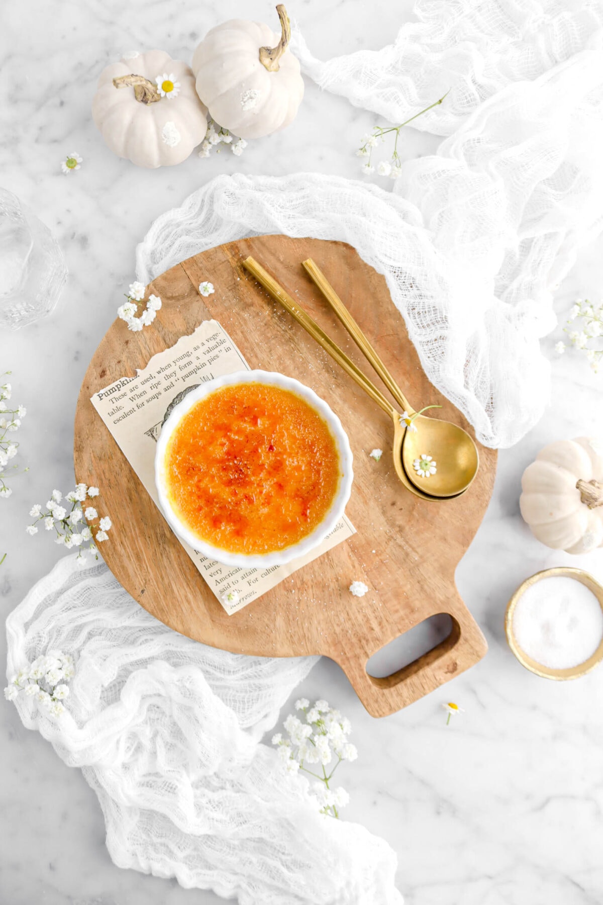 creme brûlée on wood board with book page and two gold spoons beside, with white cheesecloth, and white pumpkins on marble surface.