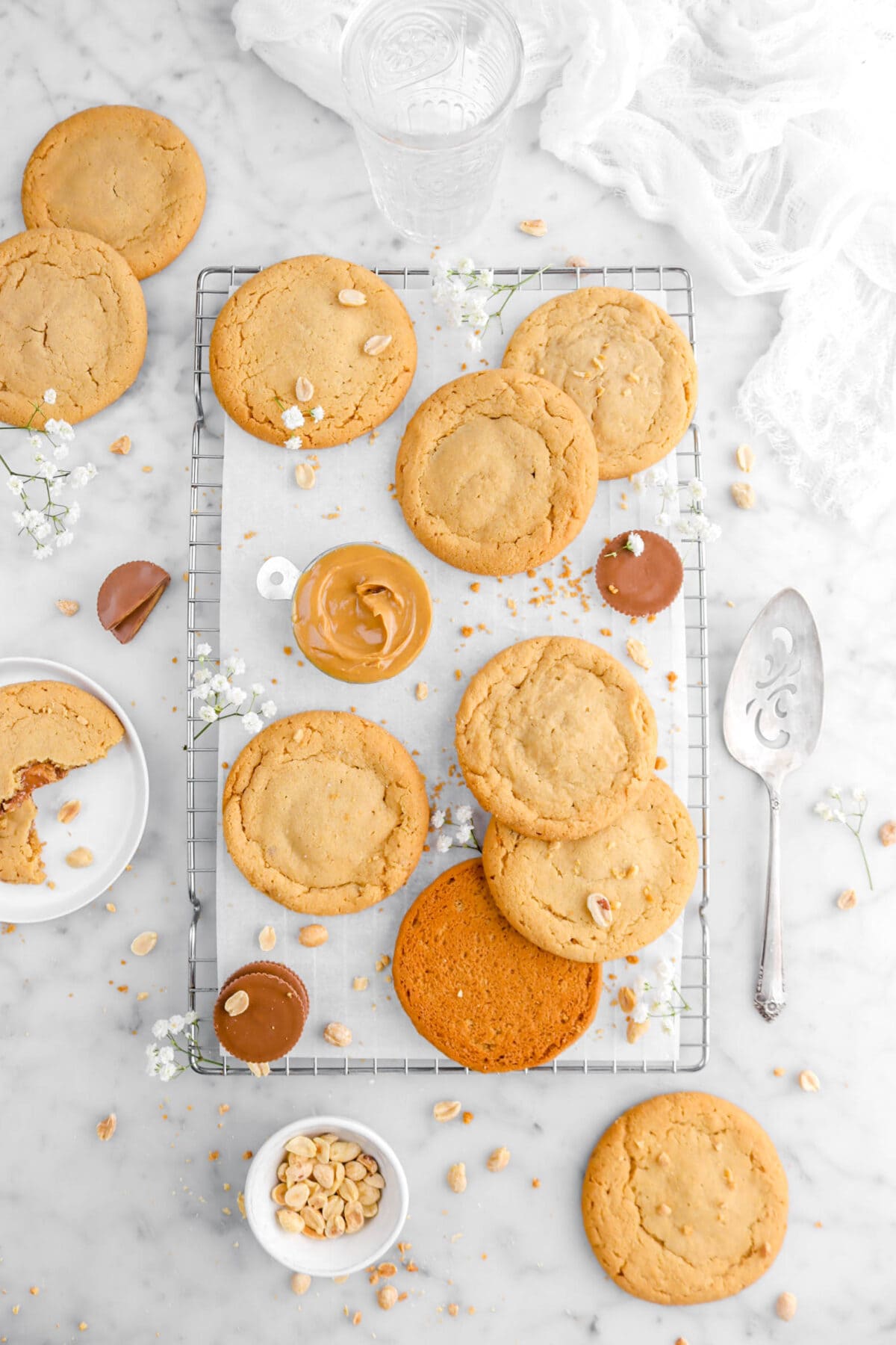 pulled back overhead shot of seven peanut butter cookies on cooling rack with measuring cup of peanut butter, peanut butte cups, peanuts, and white flowers on cooling rack with more cookies around, plate of broken in half cookie beside, and cake knife on marble surface.