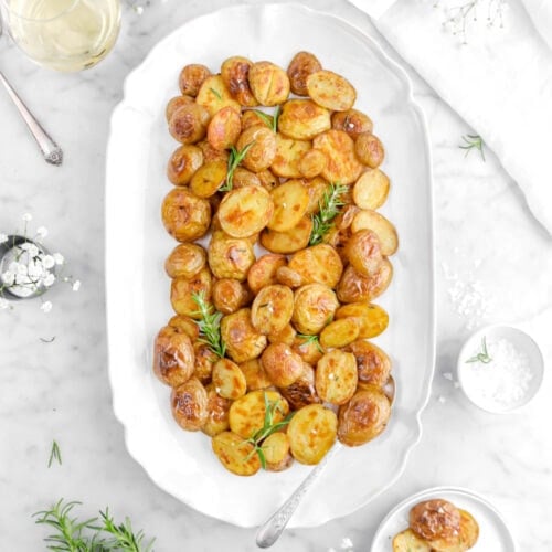 roasted potatoes on white platter with spoon and fresh rosemary sprigs.