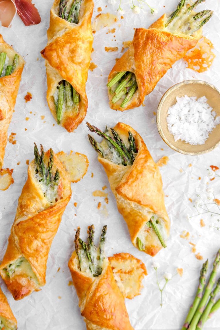 Asparagus pastry bundles on parchment paper with pastry crumbs around and a small gold bowl of salt beside