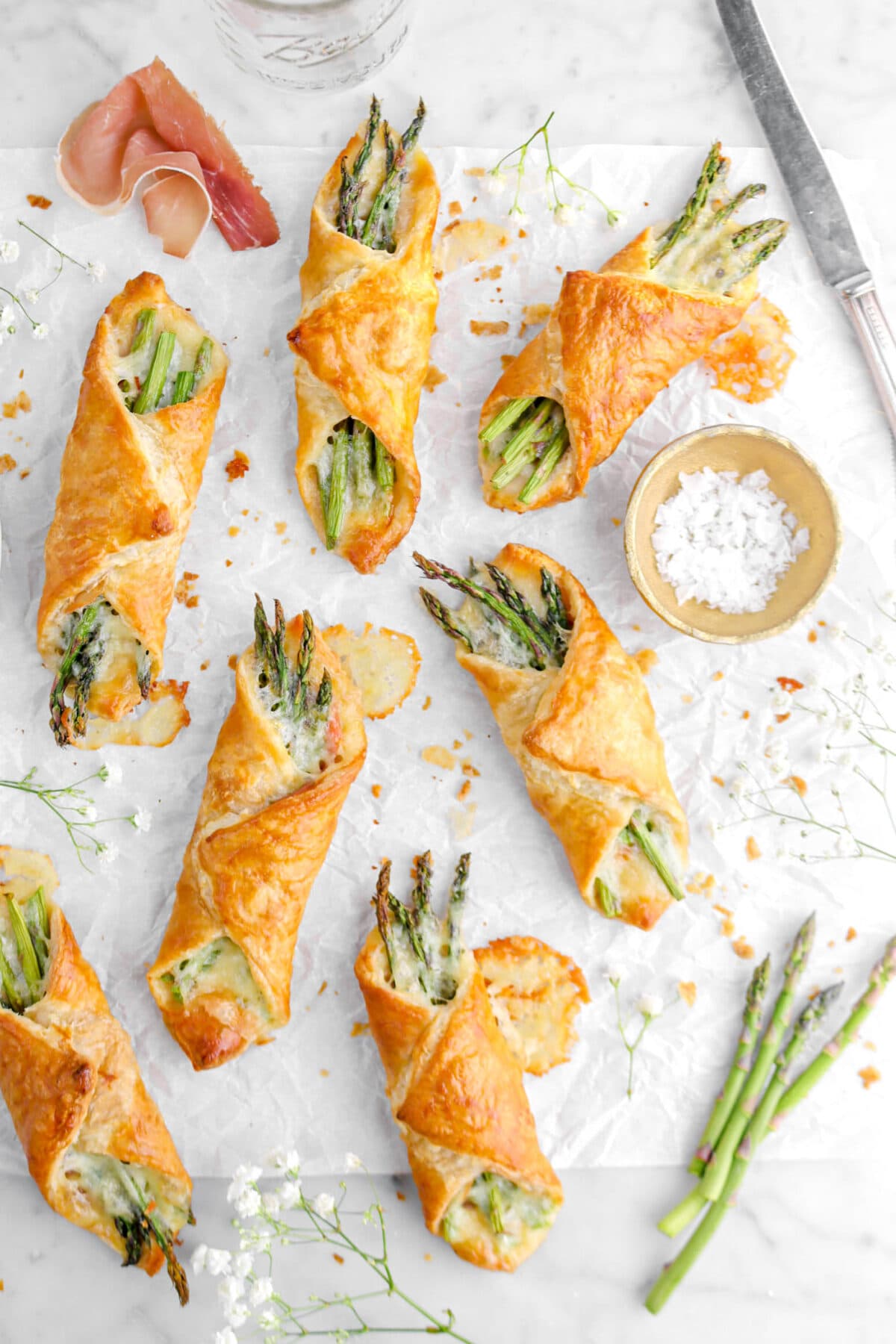 seven pastry bundles on parchment paper with melted cheese, pastry crumbs, flowers, a knife, a piece of prosciutto, and fresh asparagus.